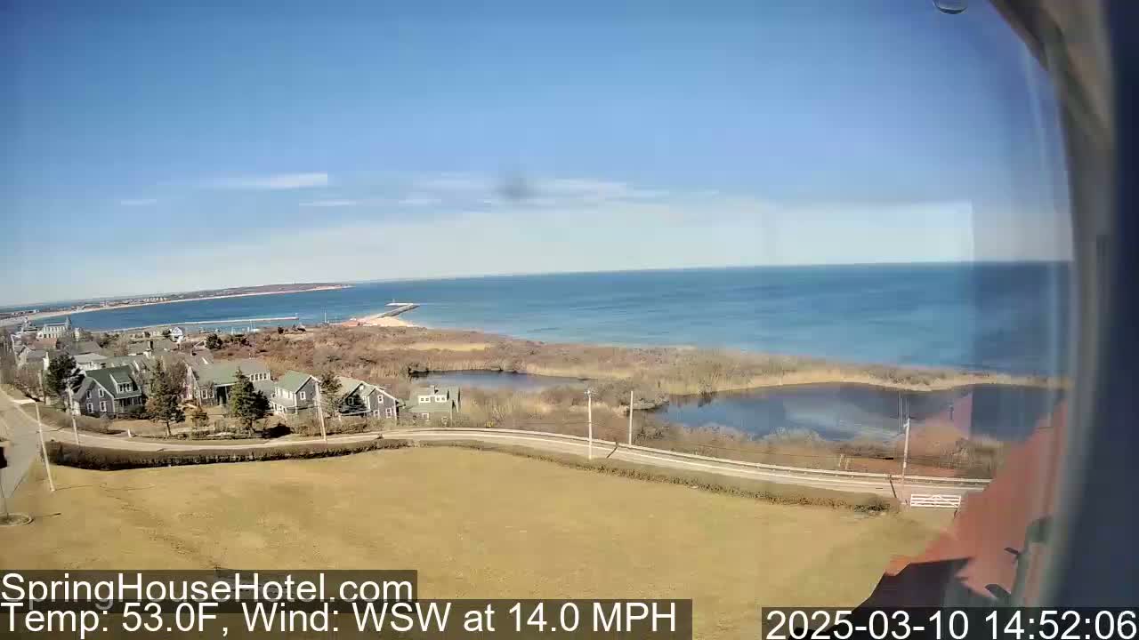 A coastal scene on a clear, sunny day with a 14 mph wind shows houses, marshland, and a calm ocean with a distant peninsula.