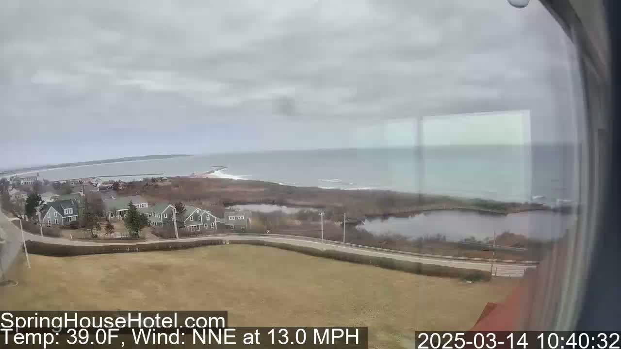 A coastal view on an overcast day shows a calm ocean, a road curving past several houses and a small pond, with a light wind blowing from the north-northeast at 13 miles per hour.