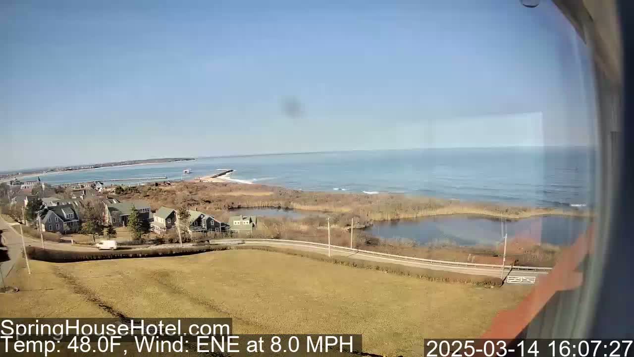 An elevated view shows a coastal scene with houses, a road, marshland, and a calm ocean under a clear blue sky.
