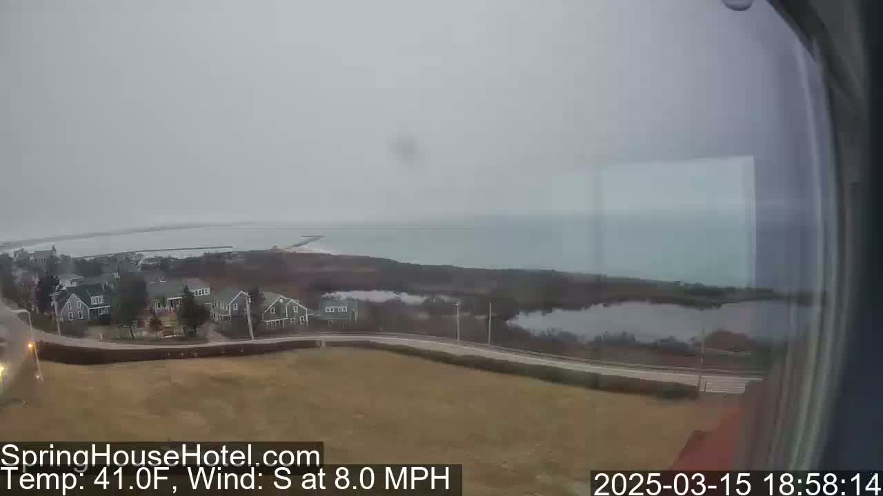 A coastal view on an overcast day shows houses, a road, a body of water, and a shoreline under a gray sky.