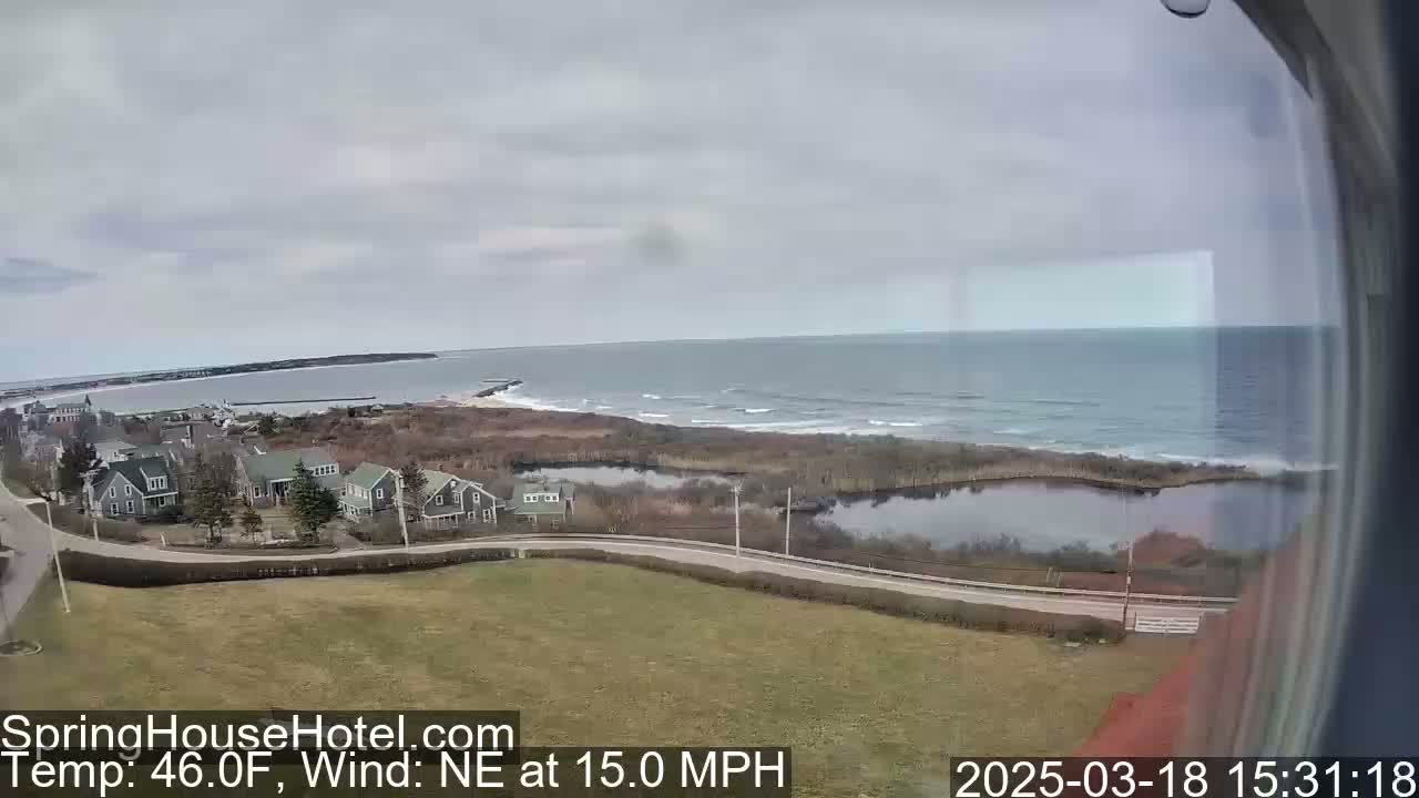 An elevated view shows a coastal scene with houses, a road, a pond, and a calm ocean under an overcast sky.