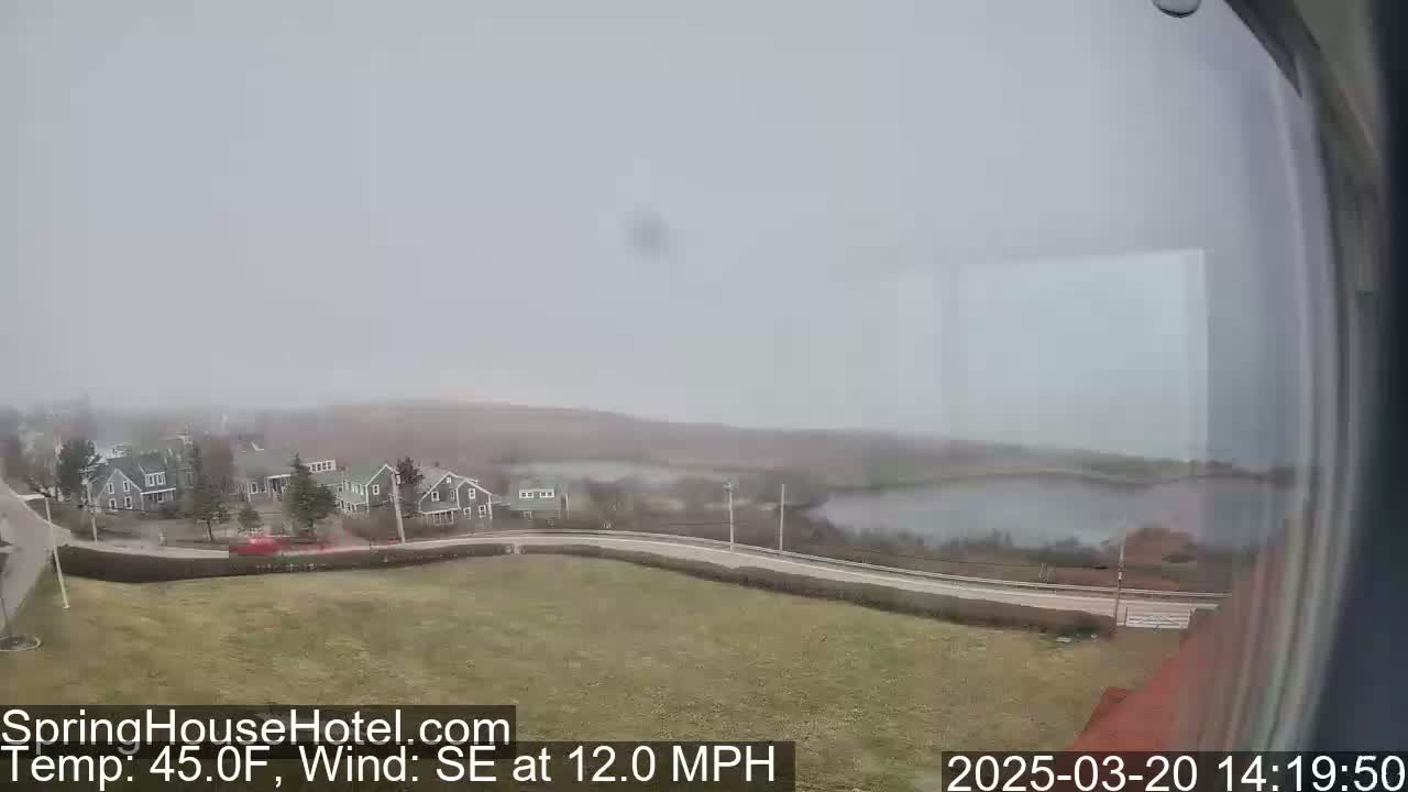 A hazy, overcast view shows a road curving past houses and a body of water, with a 12 mph southeast wind.