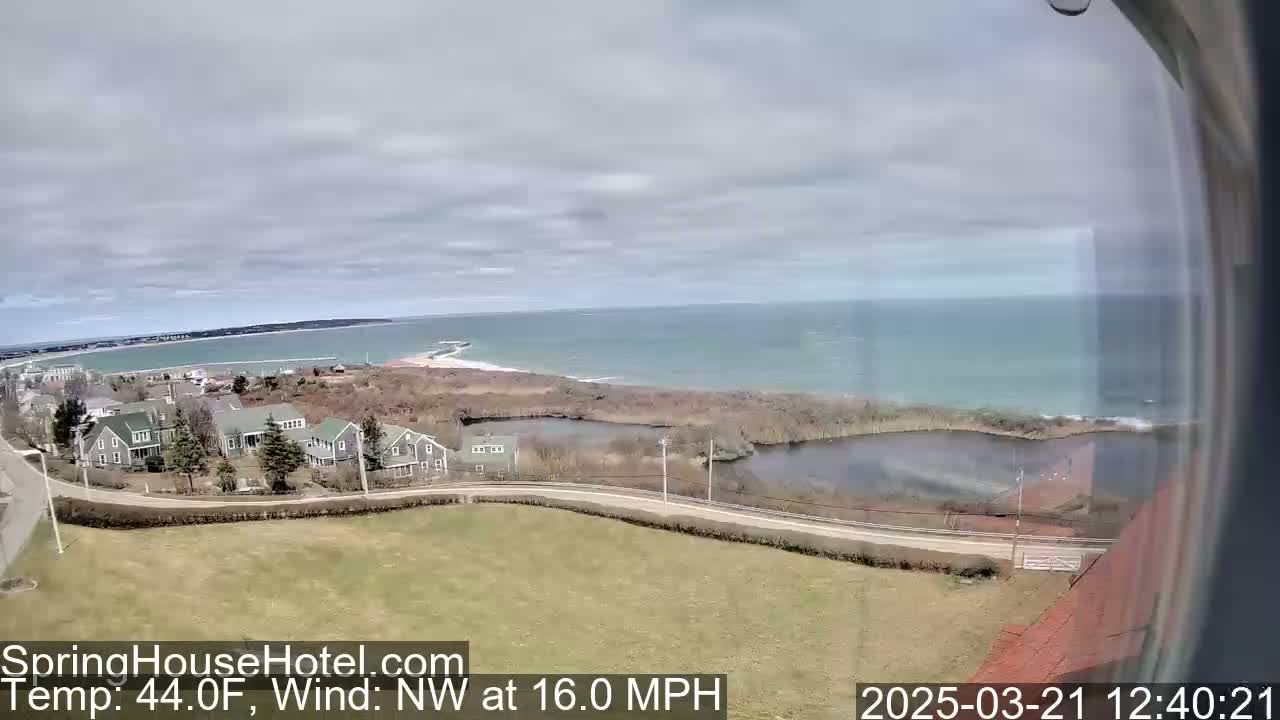 A coastal scene on a partly cloudy day shows a calm ocean, houses, and a grassy area with a road, under a northwest wind blowing at 16 miles per hour and a temperature of 44 degrees Fahrenheit.
