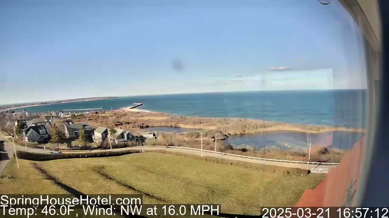 A coastal scene on a clear, sunny day with a northwest wind at 16 mph shows houses near a body of water with a shoreline extending to a calm ocean.