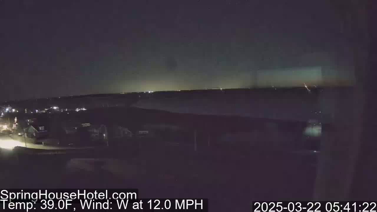 A nighttime, high-angle view shows a suburban area with a body of water in the distance under a dark sky with a 39°F temperature and a 12 mph west wind.