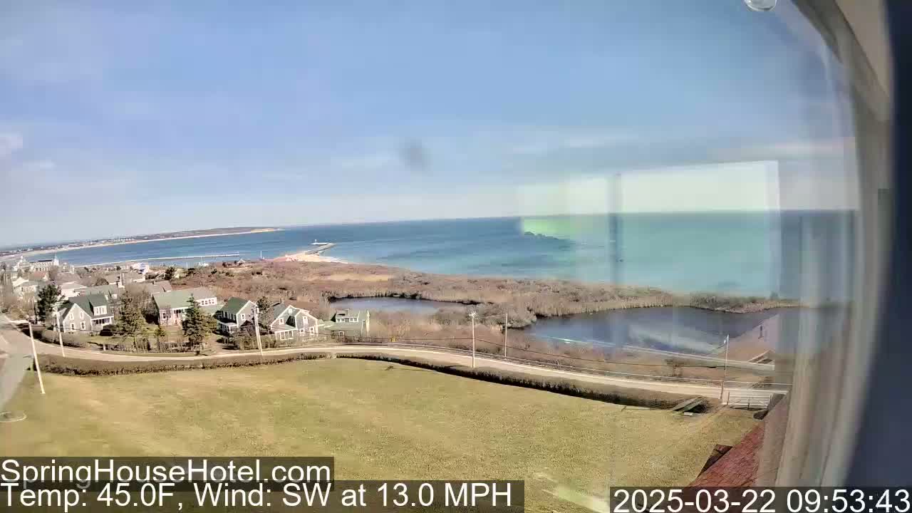 A coastal scene on a sunny day with a southwest wind of 13 mph shows a residential area overlooking a calm body of water and a distant coastline.