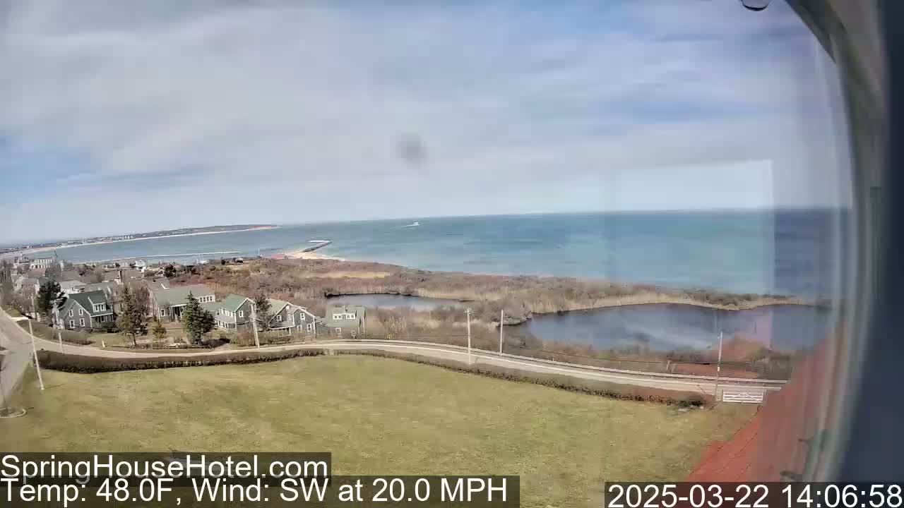 A coastal scene with houses, a road, a body of water, and a calm ocean under a mostly clear sky, with a southwest wind at 20 mph.