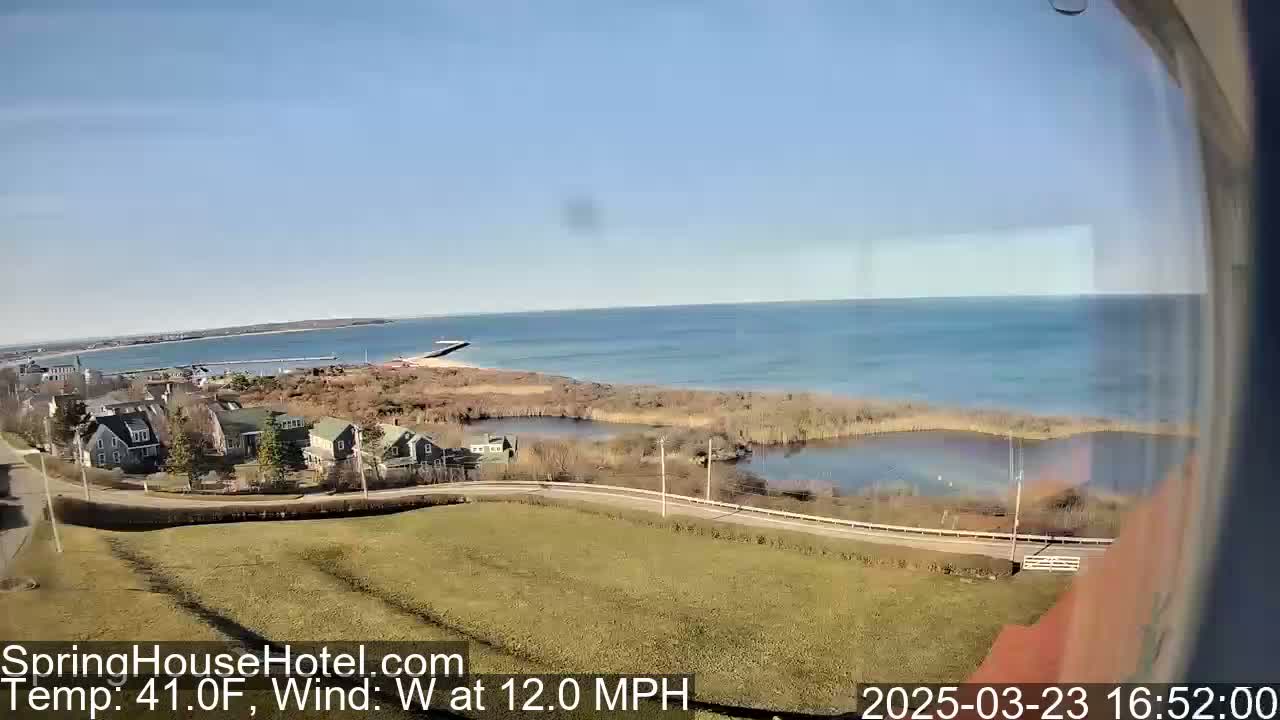 A coastal scene on a partly sunny day with a 12 mph westerly wind shows a road curving past houses near a calm body of water, which leads to a larger bay and a distant shoreline.