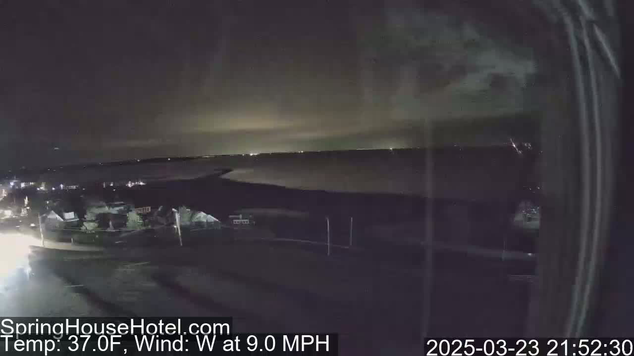 A nighttime view of a calm body of water, with a town illuminated in the foreground, under a dark sky with a slight glow on the horizon and a 9 mph westerly wind at 37 degrees Fahrenheit.