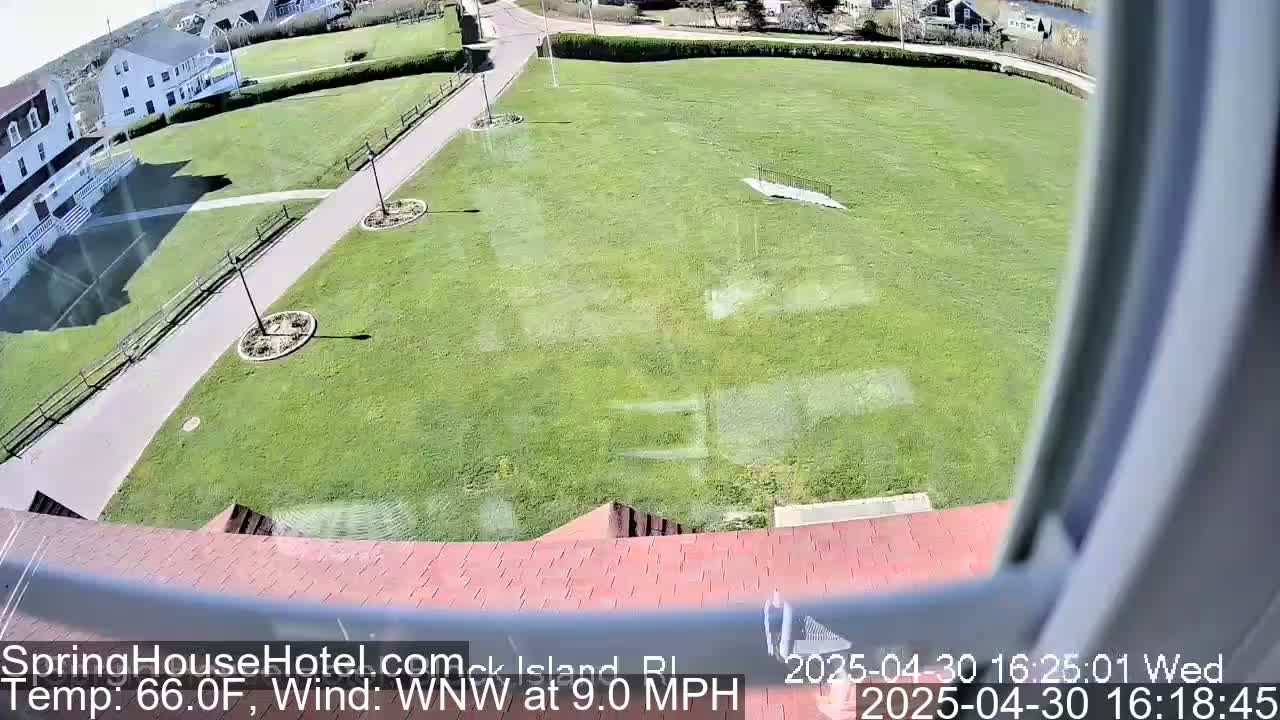 A high-angle view shows a grassy area with a paved pathway, some landscaping, and buildings in the background on a sunny day with a WNW wind at 9.0 mph.