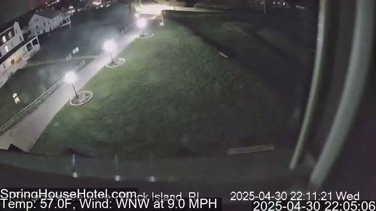 A nighttime, overhead view of a grassy area with a paved walkway, lit by several lamps, adjacent to buildings.