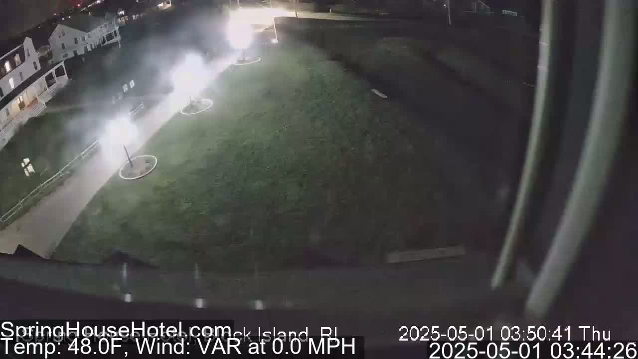 A nighttime, high-angle view shows a grassy area with several bright lights and smoke-like plumes emanating from them near a building.