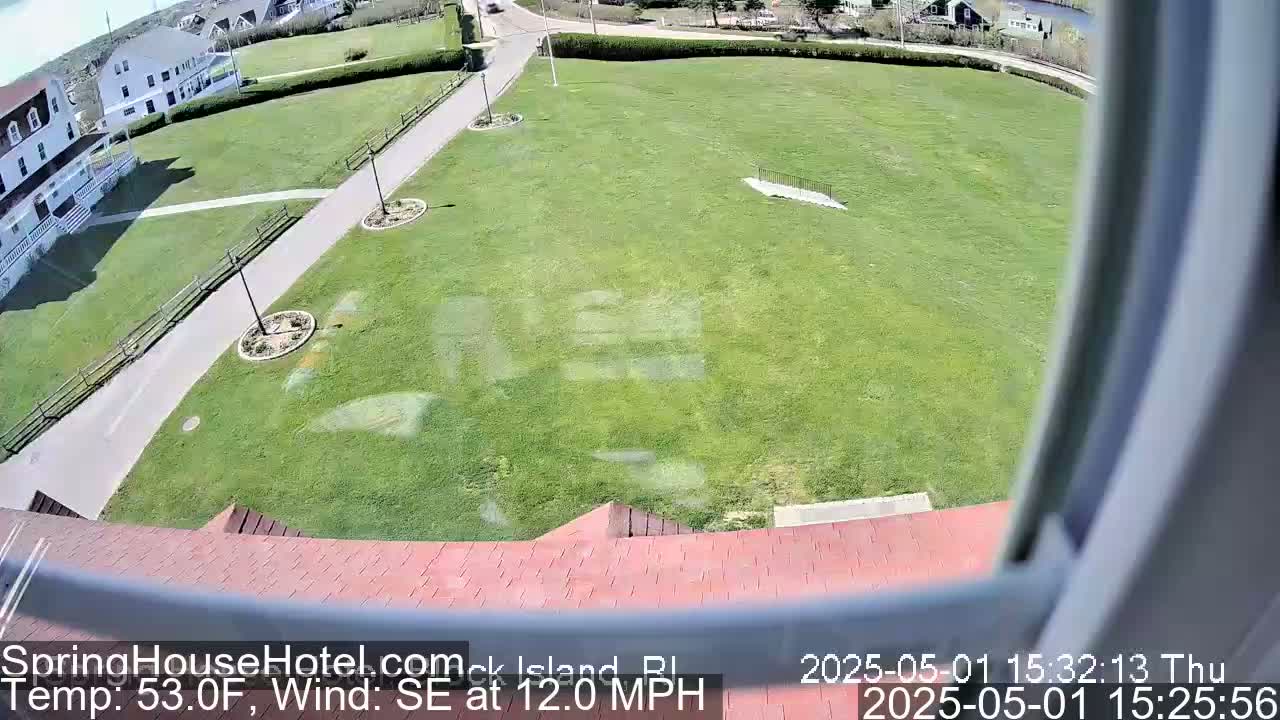 A high-angle view shows a large grassy area with a paved path and some buildings in the background, under a sunny sky with a southeast wind.