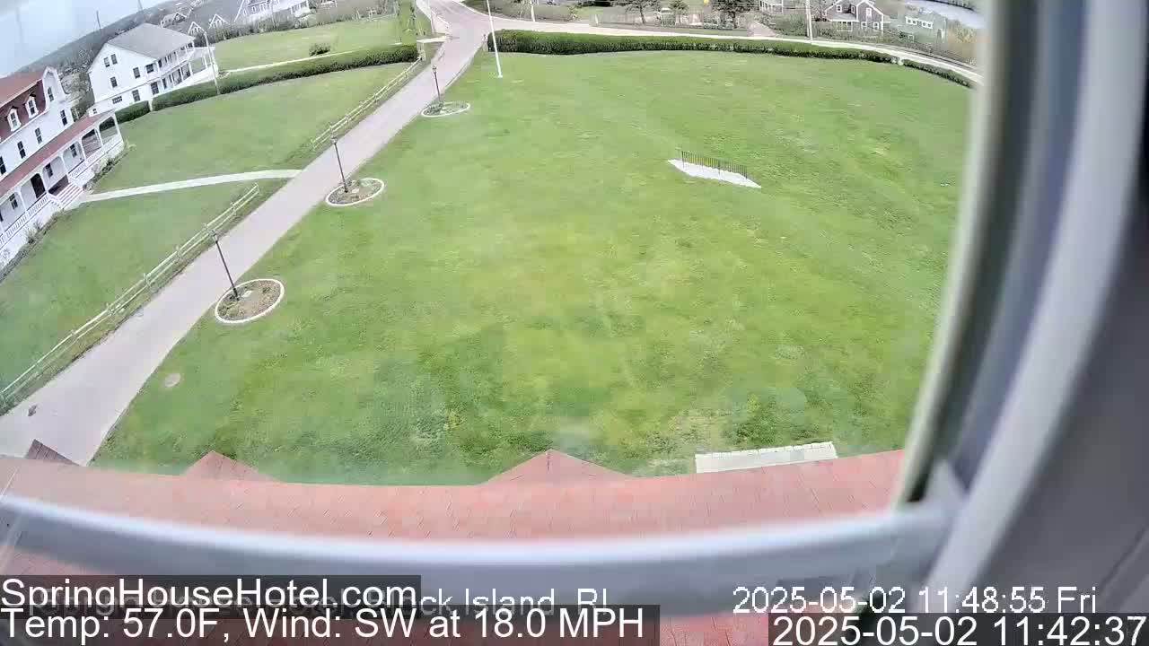 A high-angle view shows a grassy area with a paved path leading to white buildings, under a partly cloudy sky with a southwest wind at 18 mph.