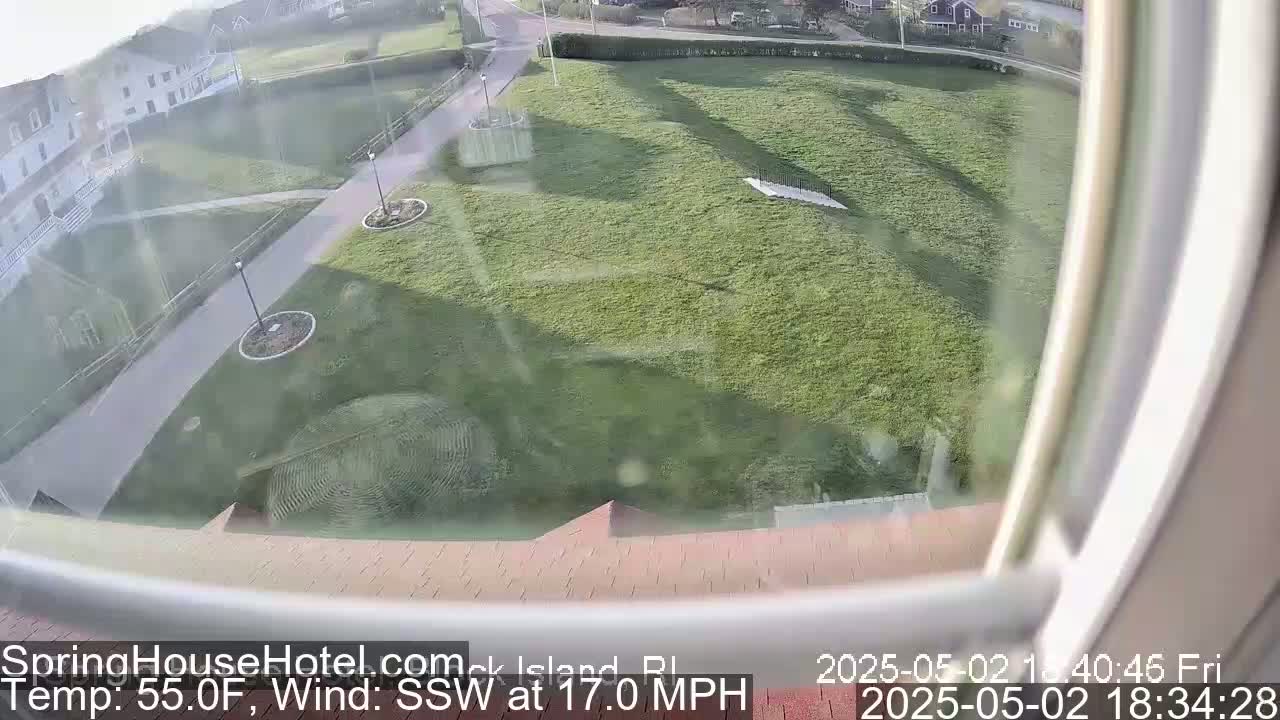 A view from a window shows a grassy area with a walkway and buildings in the background, under windy, 55°F weather.