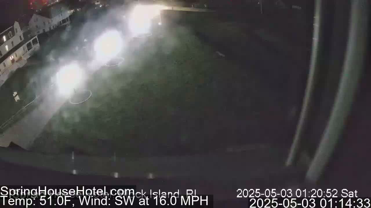 Several fireworks explode over a grassy field next to a building at night, with a southwest wind at 16 mph.