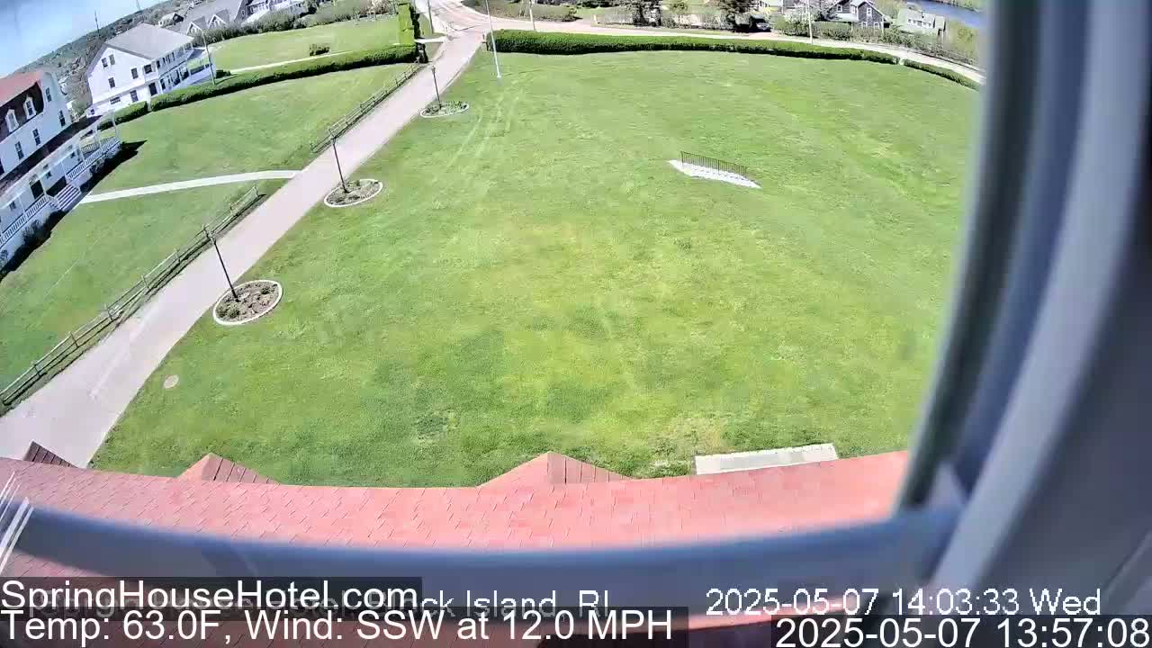 A view from a window shows a large grassy area with a paved path leading to a building, under sunny conditions with a south-southwesterly wind at 12 mph.