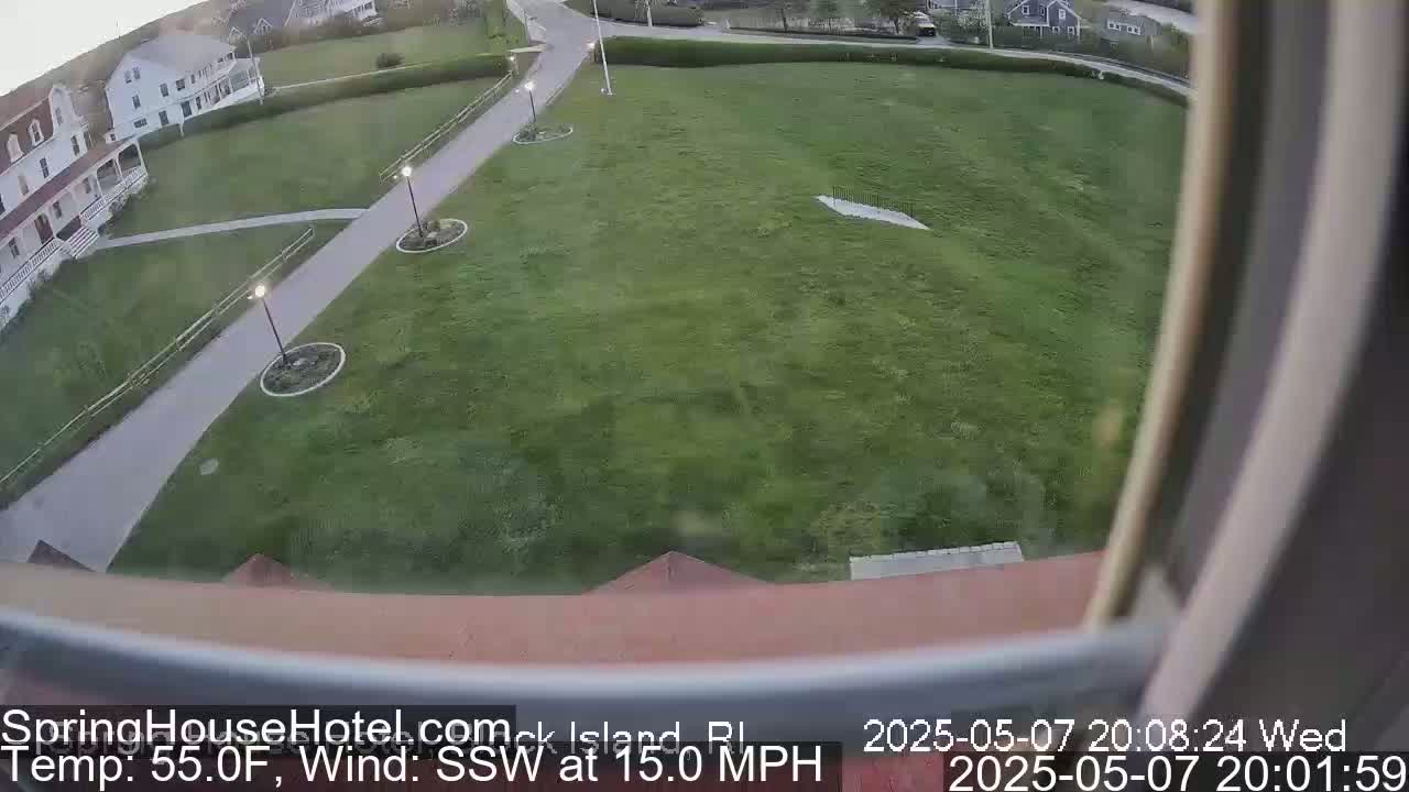 A grassy area with a paved walkway and lampposts is visible from a window, with a building partially in view, under partly cloudy conditions and a 15 mph south-southwesterly wind.