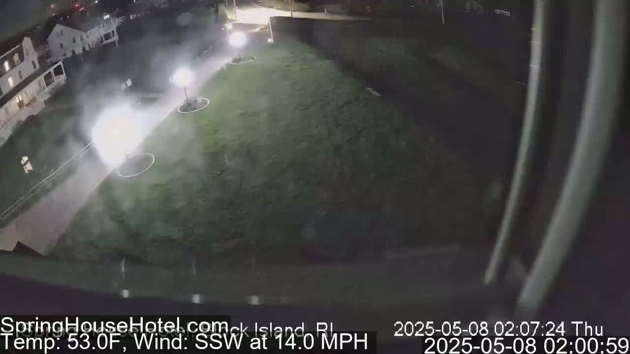 A nighttime, windy scene shows a grassy area illuminated by lights near a building, with some fog or mist present.