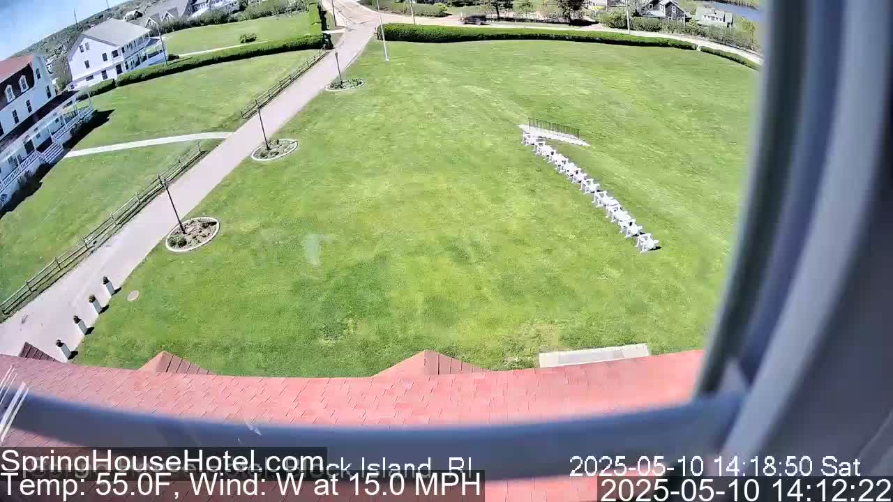 A view from a window shows a grassy lawn with a row of chairs, a building, and a paved walkway, all under sunny skies with a west wind at 15 mph.