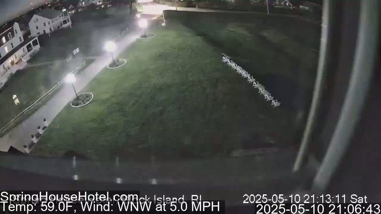 A nighttime, high-angle view shows a grassy area with several lights and a row of objects on the grass, adjacent to a paved walkway and buildings, with a slight wind.