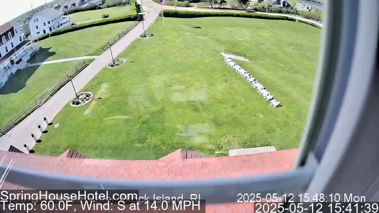 A high-angle view shows a grassy area with a row of white chairs, a paved walkway, and a building in the background on a sunny day with a south wind of 14 mph and a temperature of 60°F.