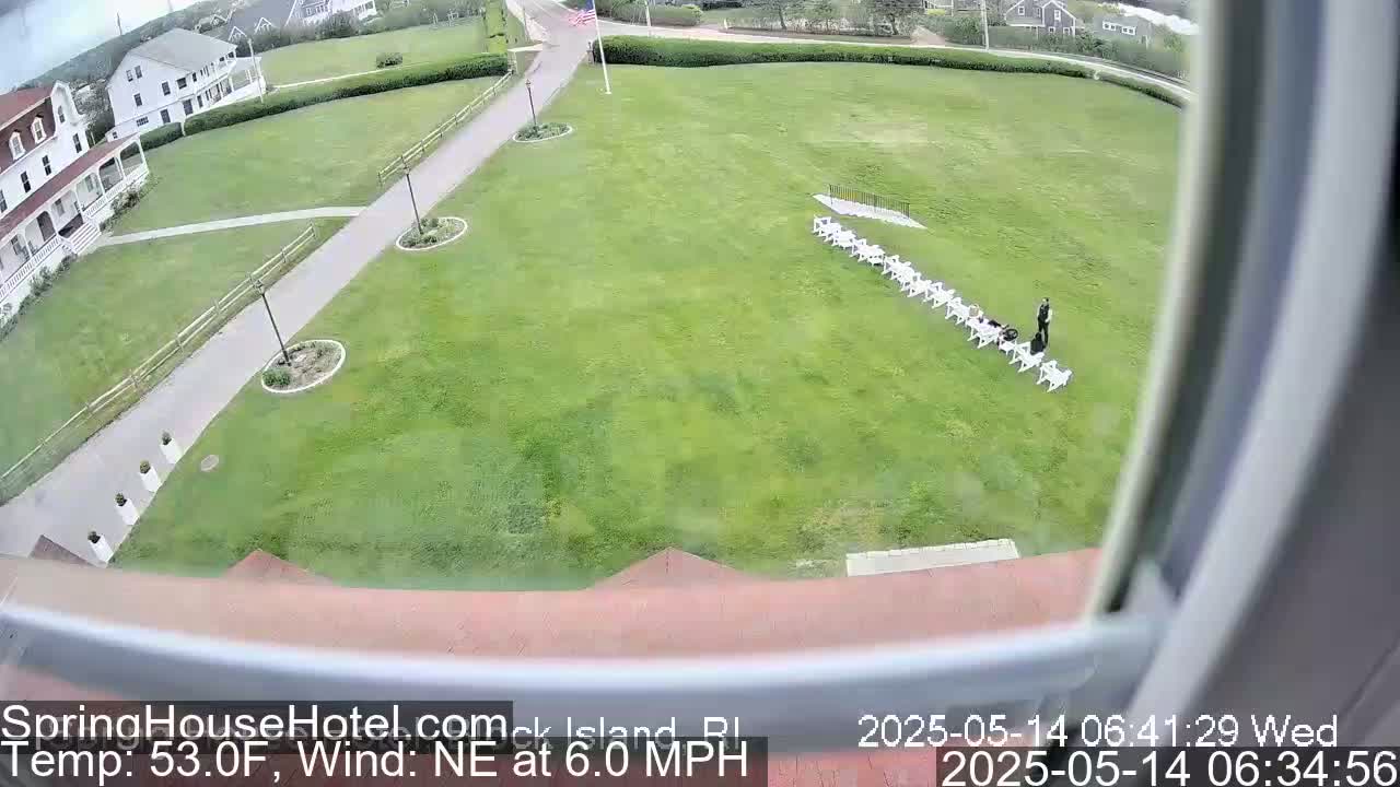 A grassy lawn with a row of white chairs and a person standing nearby is seen from an elevated viewpoint, with a building and calm weather in the background.