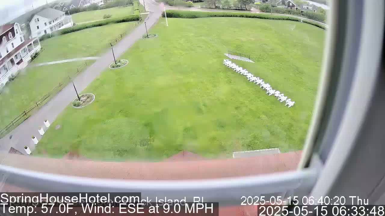 A high-angle view shows a grassy lawn with a row of white chairs, a paved path, and a white building in the background, with a slight breeze from the east-southeast.