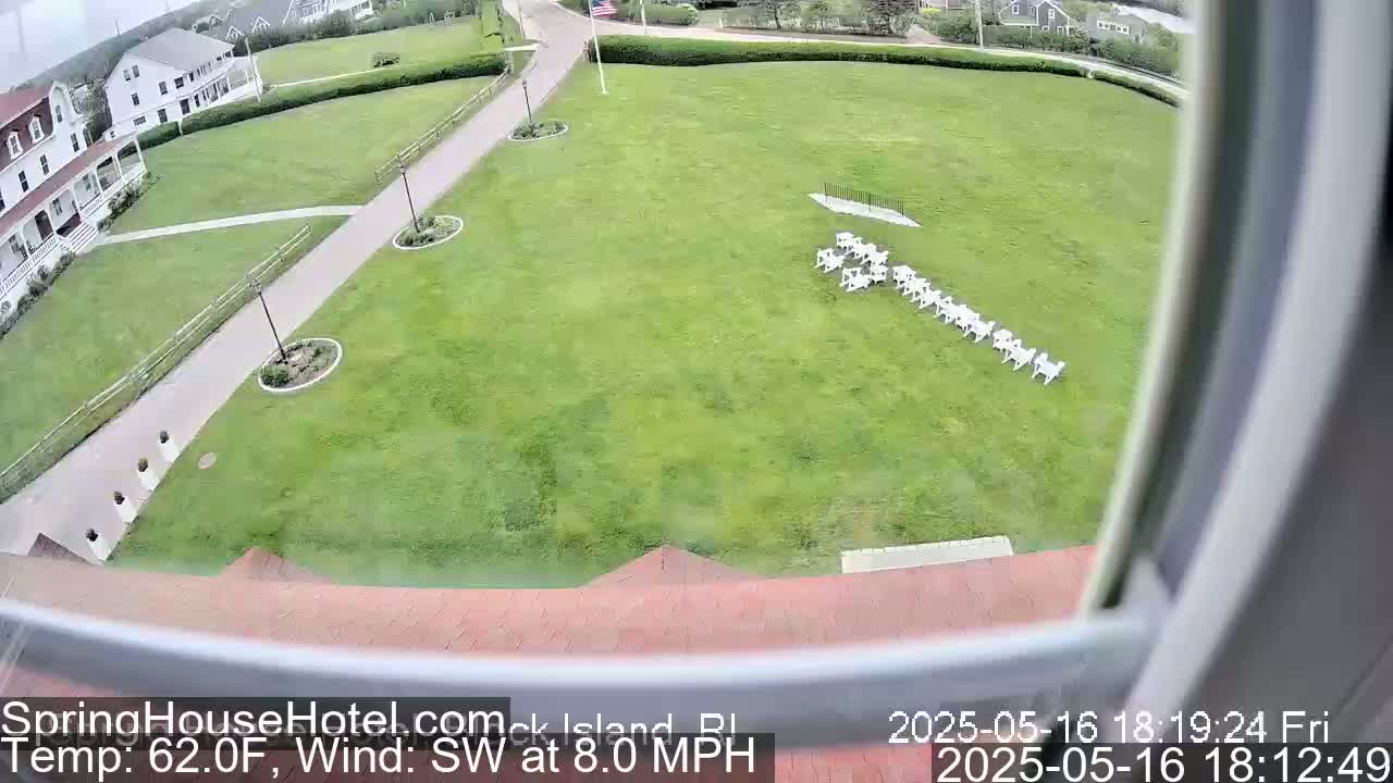 A high-angle view shows a large grassy area with a row of white chairs, a building, and paved walkways, all under a partly cloudy sky.