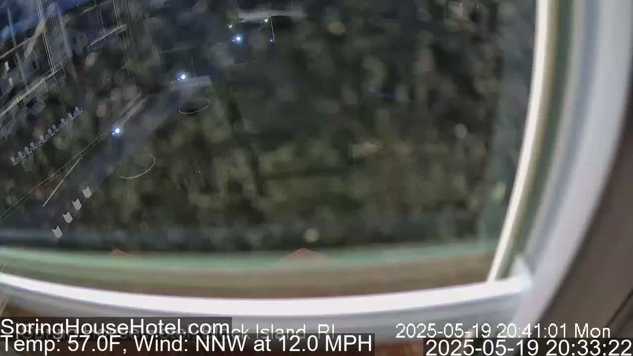 A nighttime view through a window shows a blurry scene of a road, some vegetation, and a few faint lights, with a northwest wind at 12 mph and a temperature of 57 degrees Fahrenheit.