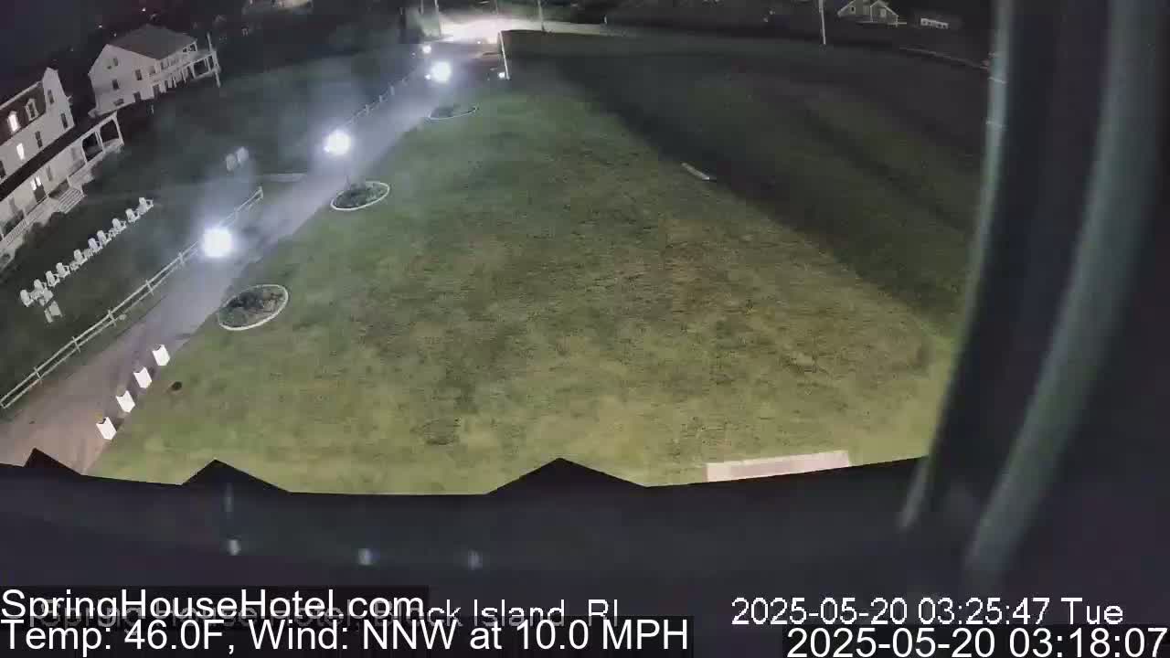 A nighttime, high-angle view of a grassy area next to a building with a paved walkway, illuminated by several lights, under a northwest wind of 10 mph and a temperature of 46°F.