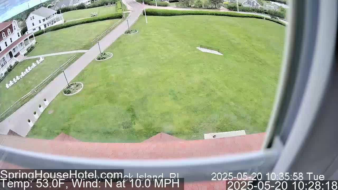 A wide shot from a window shows a large expanse of green grass, a building, and paved walkways on a partly cloudy day with a north wind at 10 mph.