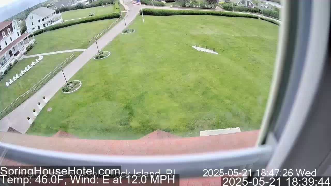 A wide shot from a window shows a large grassy area with walkways, landscaping, and a white building in the background, under partly cloudy skies with a light east wind.