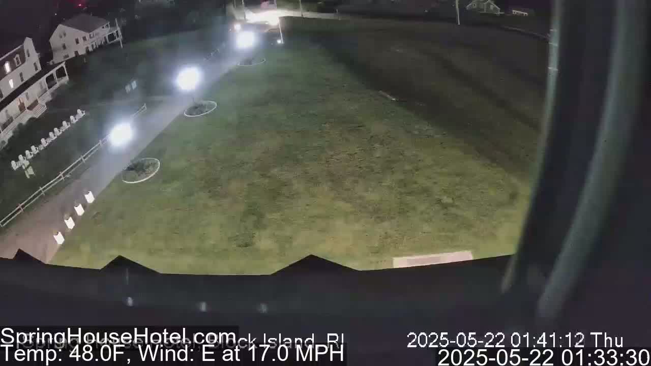 A nighttime, high-angle view shows a grassy area with a paved walkway, buildings, and outdoor lighting, under an east wind of 17 mph and a temperature of 48°F.