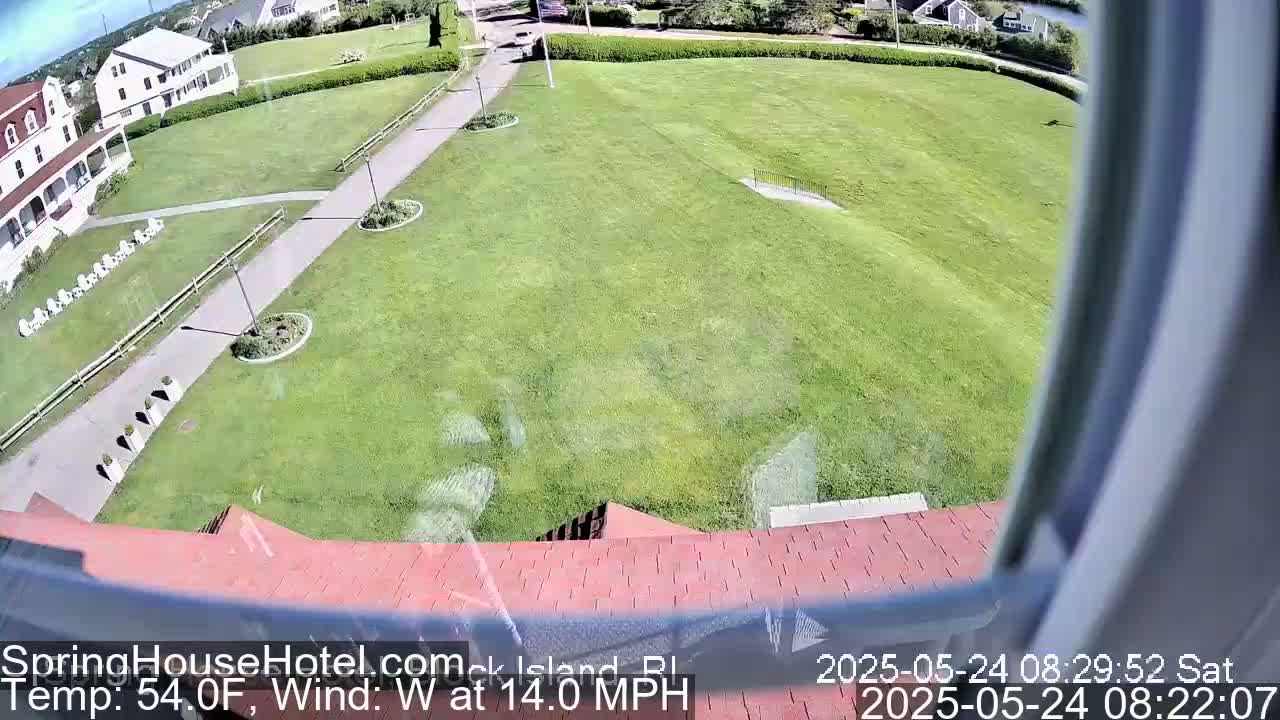 A view from a window shows a large grassy area with a paved walkway leading to a building, all under partly sunny skies with a west wind.