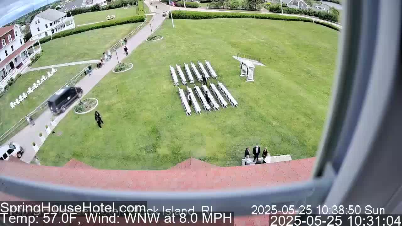 An outdoor scene on a sunny day with an 8 mph wind shows a large grassy area with rows of white chairs set up for an event, a few people walking around, and buildings in the background.