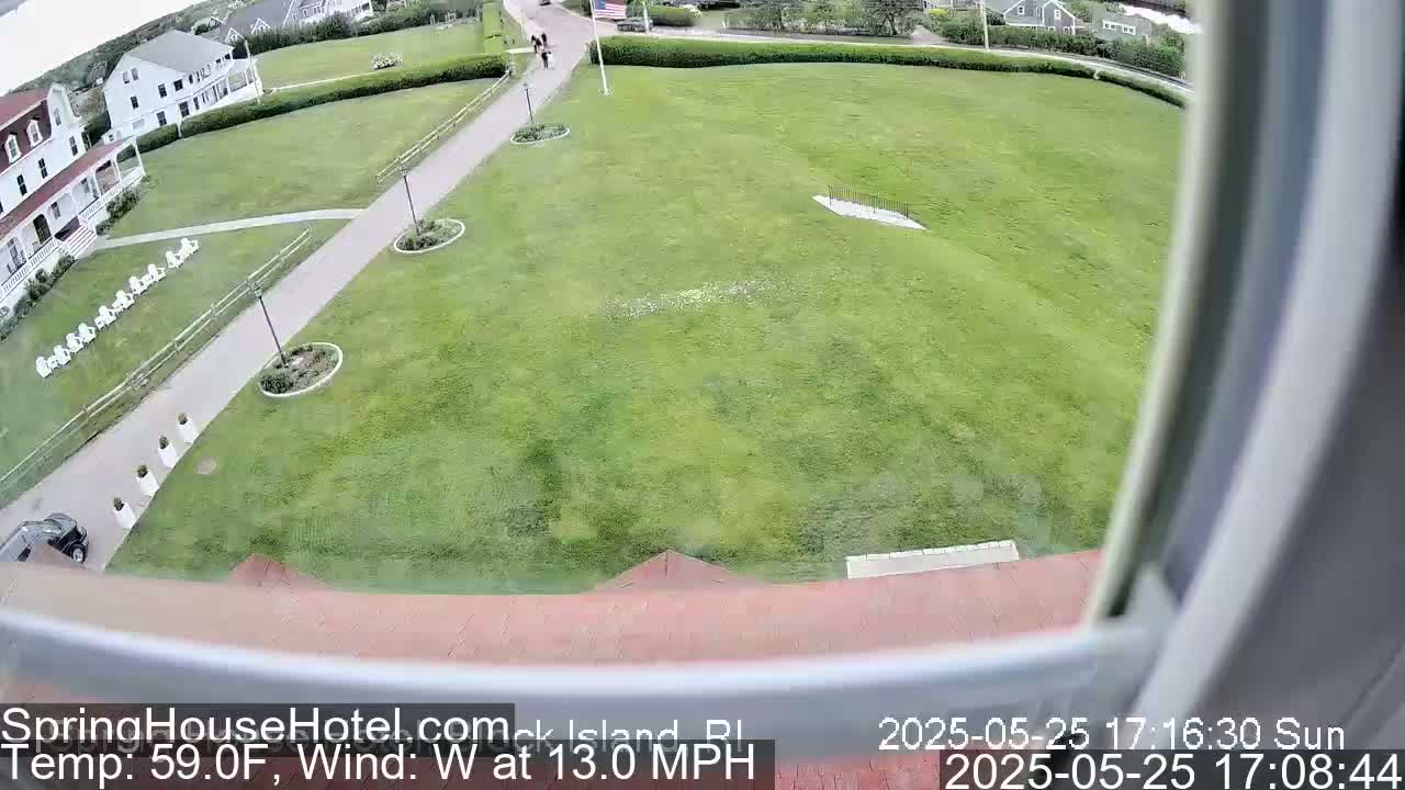 A view from a window shows a large grassy area with a building and some landscaping, under sunny conditions with a west wind at 13 mph.
