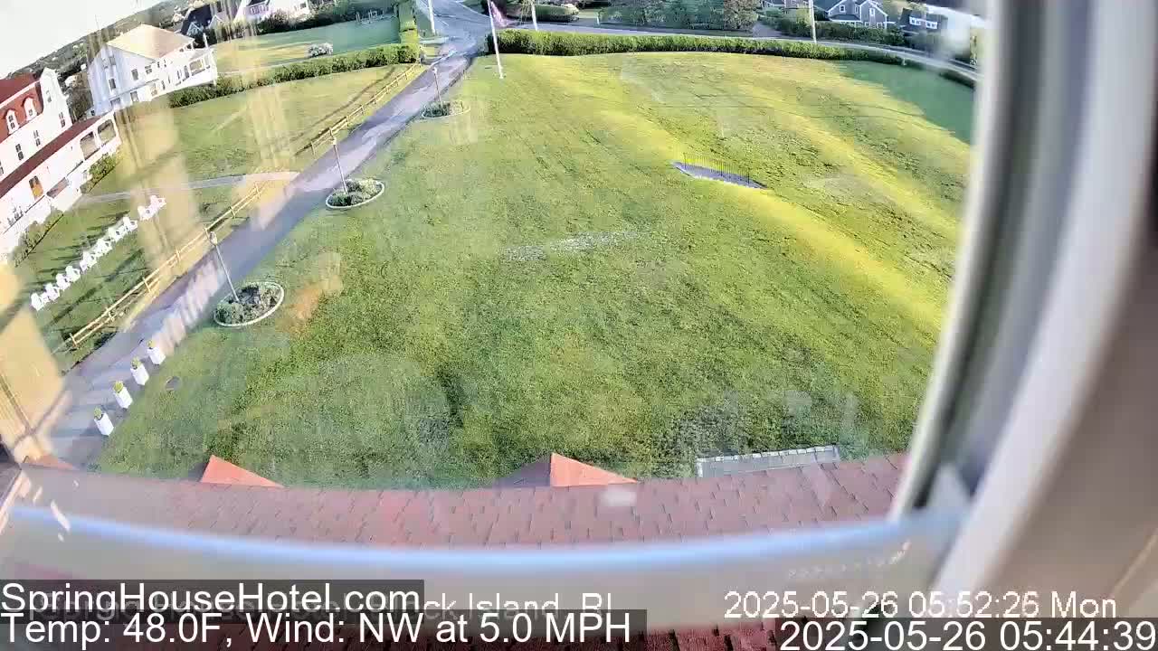 A view from a window shows a grassy area with a path, some landscaping, and a building in the background, under partly sunny conditions with a northwest wind.