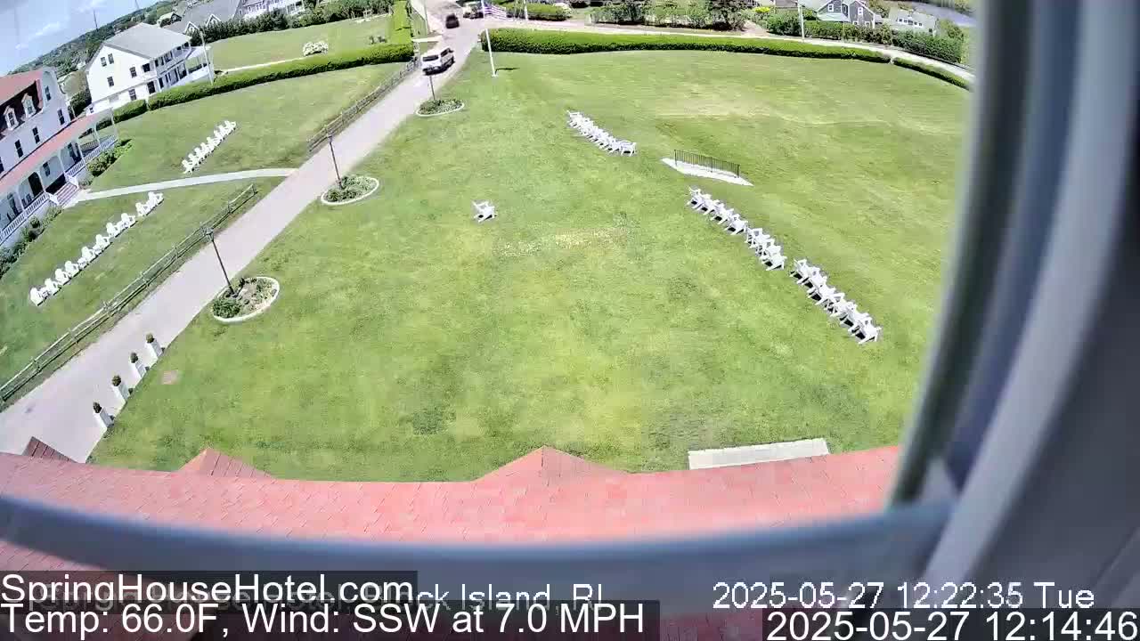 A high-angle view shows a large grassy area with rows of white chairs, a building with a red roof, and a paved pathway, all under sunny conditions with a southwest wind at 7 mph.