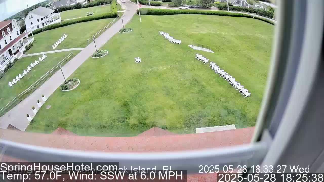 A large grassy area with rows of white chairs is seen from an elevated vantage point, with a building and paved walkways visible in the background on a partly sunny day with a south-southwesterly wind of 6 mph.