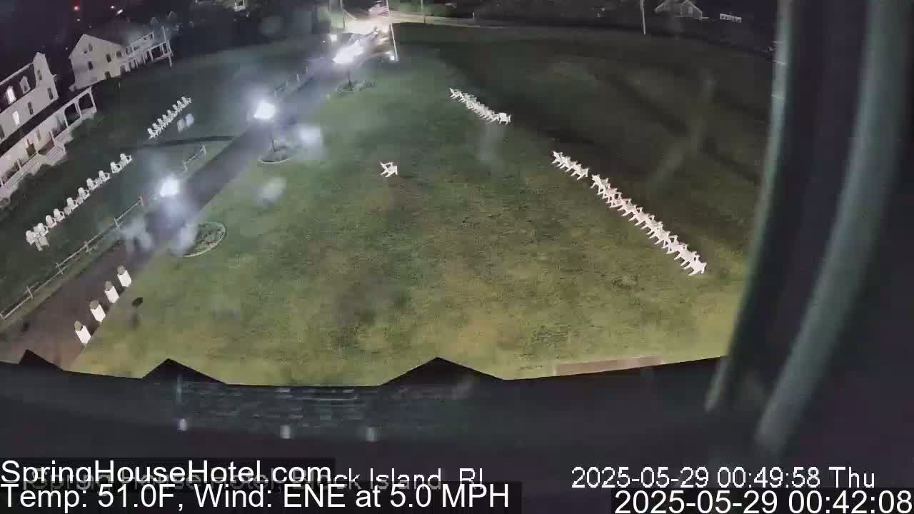 A nighttime aerial view shows a grassy area with rows of white chairs, a building, and a paved walkway, under a clear sky with a 5 mph east-northeast wind and a temperature of 51 degrees Fahrenheit.