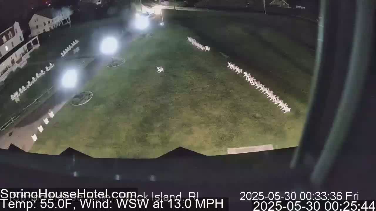 At night, under a WSW wind of 13 mph and a temperature of 55°F, rows of white chairs are illuminated on a grassy lawn next to a building.