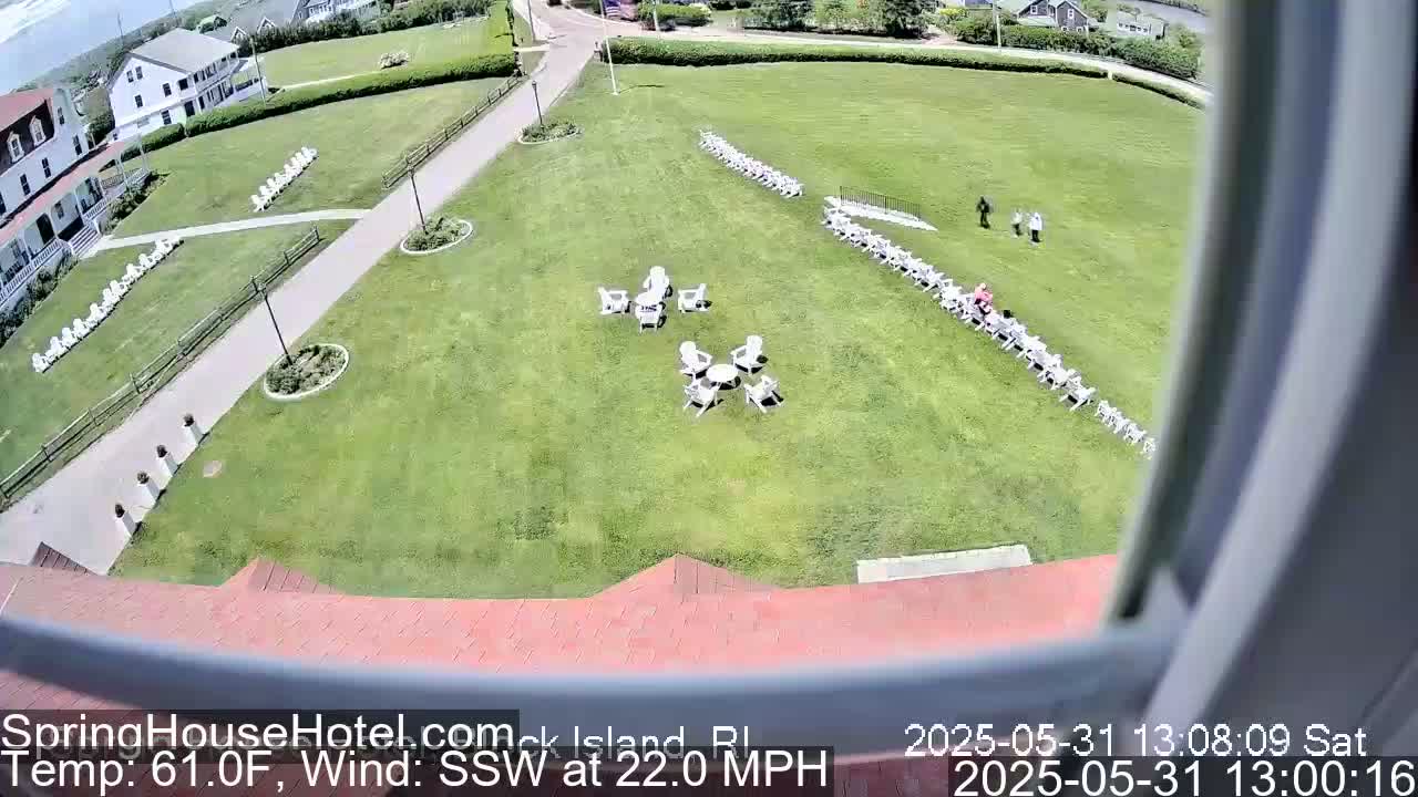 A high-angle view shows a grassy lawn with several rows of white chairs, small groups of chairs around tables, and a few people walking, all under a sunny sky with a southwest wind at 22 mph and a temperature of 61 degrees Fahrenheit.