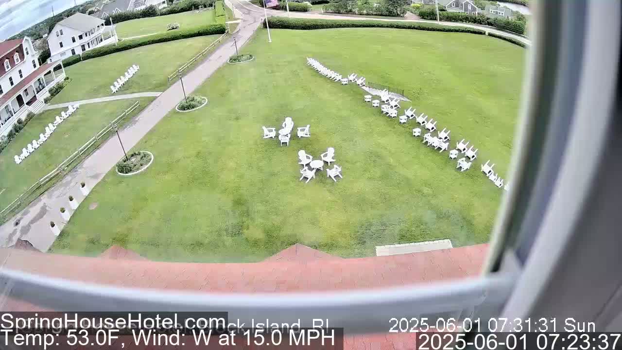 A grassy lawn with numerous white chairs arranged in groups and lines, seen from an elevated viewpoint on a sunny day with a west wind of 15 mph and a temperature of 53 degrees Fahrenheit.