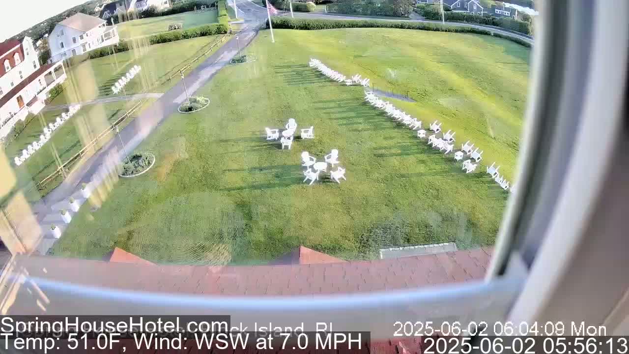 A grassy area with numerous white chairs arranged in rows and small groups, viewed from a window on a partly sunny day with a WSW wind at 7.0 mph and a temperature of 51.0°F.