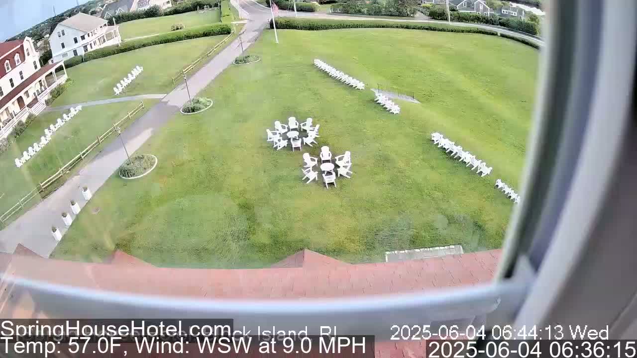 A grassy lawn with numerous white chairs arranged in rows and small groups around tables, viewed from above on a partly sunny day with a wind of 9.0 mph from the west-southwest.