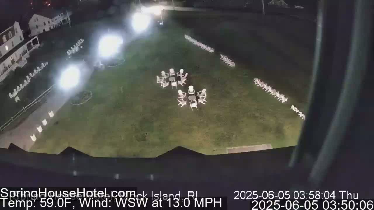 A nighttime, overhead view shows a grassy area with numerous white chairs arranged in rows and small circles, illuminated by bright lights, with a building visible in the background under a clear sky.