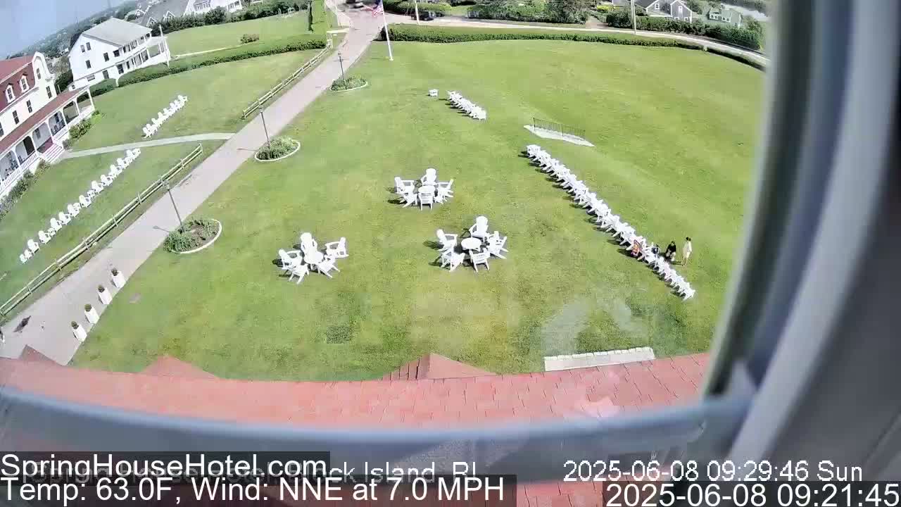 A sunny day reveals a large grassy area with numerous white chairs and tables arranged in groups,  adjacent to a building and walkway.