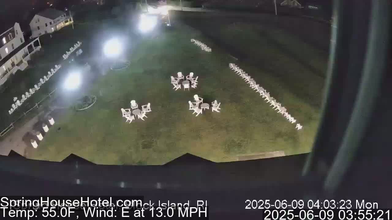 An aerial nighttime view shows a grassy area with numerous white chairs arranged in rows and small groups around tables, illuminated by artificial lights under an east wind at 13 mph and a temperature of 55°F.
