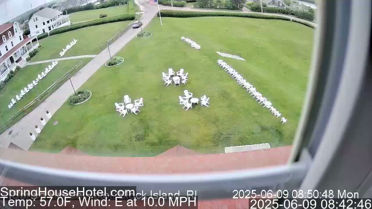 A large grassy area with several sets of white outdoor chairs and tables, some arranged in circles, is visible from a window, with a building and a road in the background under partly cloudy skies.